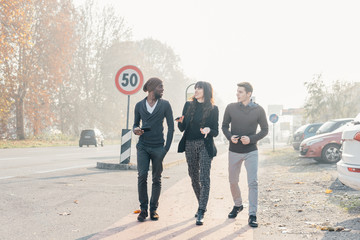 Three multiethnic woman and men friends walking outdoor in city back light, chatting and holding...