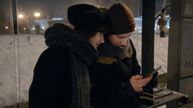 Young man and woman using touch pad on city bus station at night, information led display and people getting into the bus in background