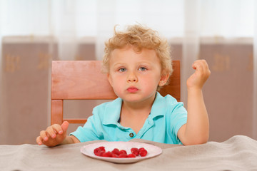 Little blonde curly-haired kid in a blue shirt had eaten red raspberries for breakfast out of pink porcelain plate, sitting at the kitchen table on a chair with a high wooden back.