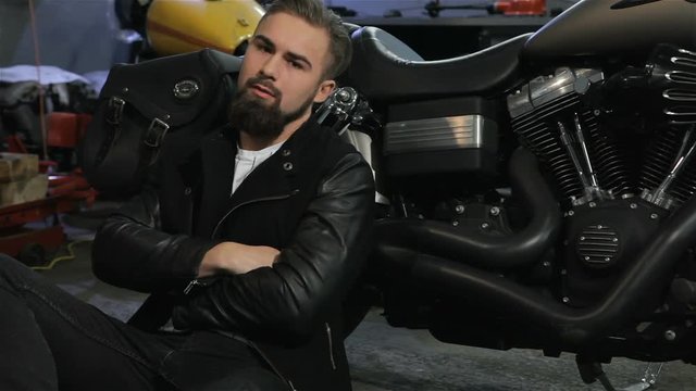 Male biker sitting on the floor near the motorcycle. Young man in black leather jacket leaning on the engine of motorcycle by his back. Bearded caucasian man holding his hands crossed on the chest