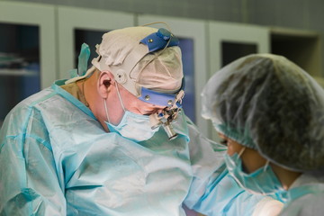 Surgeon and his assistant performing cosmetic surgery in hospital operating room. Surgeon in a mask wearing surgical loupes. Breast augmentation at the clinic. Mammaplasty