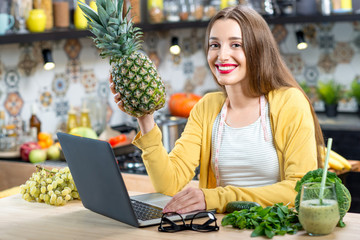 Young woman ordering healthy food online with laptop in the kitchen full with fruits and vegetables
