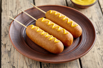 Corn dog street junk food fried hotdog meat sausage snack with mustard snack treat coated in a thick layer of cornmeal batter on stick unhealthy eating on rustic wooden desk.