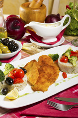 Portion, two pieces of deep fried fish rolled in bread crumbs and corn flour; side dish of boiled rice and broccoli; fresh cherry tomatoes, olives and lemon.