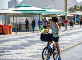 Boy with curly hair with his head turned back standing with bicycle and with dog in a basket on the bike path of Copacabana beach with the background of blurry walking people, Rio de Janeiro, Brazil