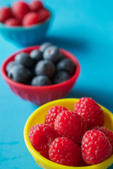 Berries in colorful bowls lined up on blue background, viewed from an angle