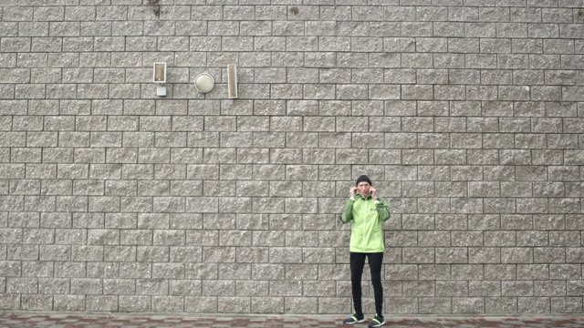 Wide shot of senior man standing against brick wall putting earphones on and then starting running