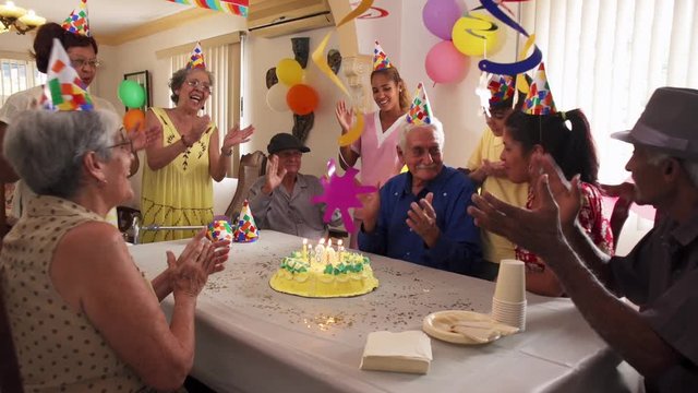 Group of old friends and family celebrating senior man birthday in retirement home. Happy elderly people having fun during party. Grandfather blowing candles on cake and smiling. Slow motion