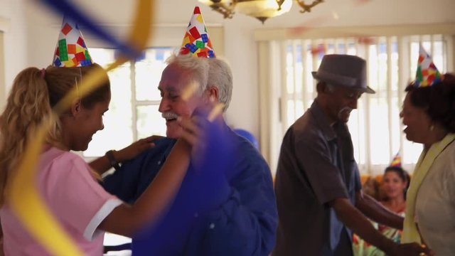 Group of old friends and family celebrating senior man birthday in retirement home. Happy elderly people having fun during party. Patient dancing with nurse in hospice and smiling. Slow motion
