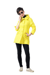 Serious short hair woman in yellow coat talking on the phone looking down. Full body length portrait isolated over gray studio background.