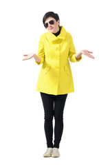 Confused woman in yellow coat wearing sunglasses shrugging shoulders. Full body length portrait isolated over gray studio background.