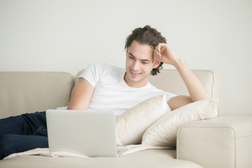 Young handsome carefree brunette man working on laptop, lifting up his spirits by listening to music via laptop, couch-potato, chatting with a long distance friend, playing online games, starting blog