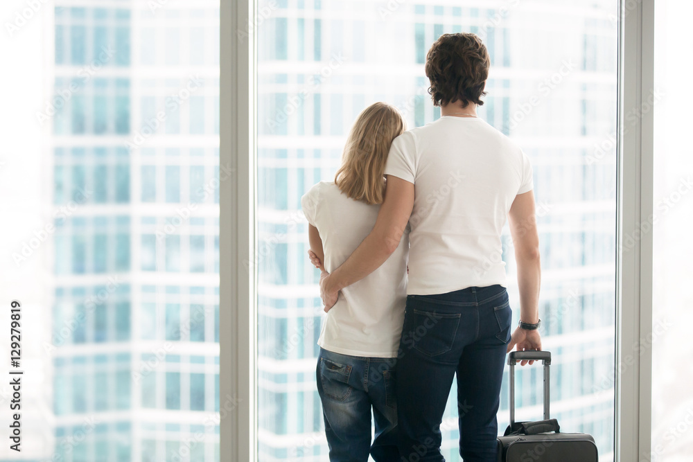 Wall mural Rear view of attractive young couple in modern apartment looking in full length window at big city scenery, holding suitcase, leaving hometown, or arriving to new place, travelling together. Indoors