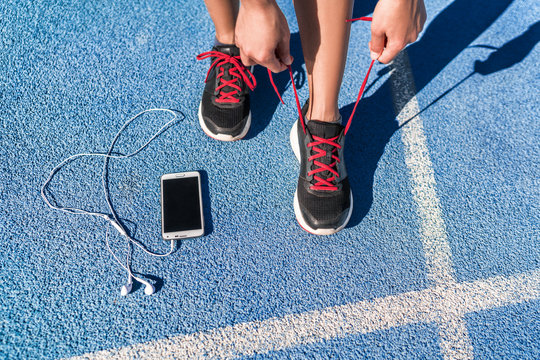 Running motivation music on mobile phone runner woman tying shoe laces. Shoes and feet closeup, athlete getting ready for race or workout on run track with smartphone and earphones.