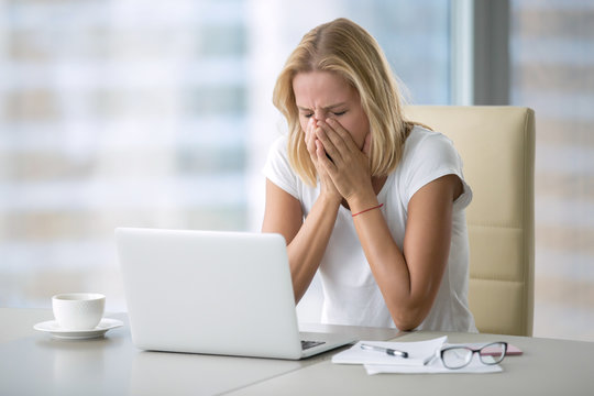 Young attractive woman at a modern office desk, with laptop covering her face with palms because of sneezing, yawning, crying. Office manners, social skills, seasonal infection, sadness concept