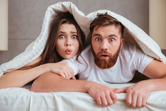 Shocked loving couple looking at camera while lying under blanket