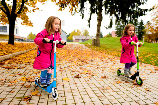 Twin girls in pink coat riding scooter on maple leaves.
