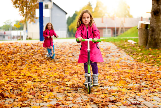 Twin girls in pink coat riding scooter on maple leaves.