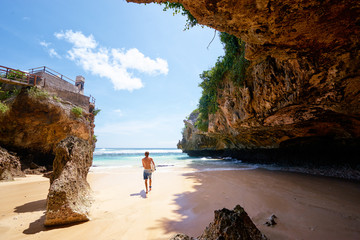 Hobby and vacation. Young man with surfboard on beautiful beach with high rocks. Uluwatu spot, Bali island, Indonesia. - obrazy, fototapety, plakaty