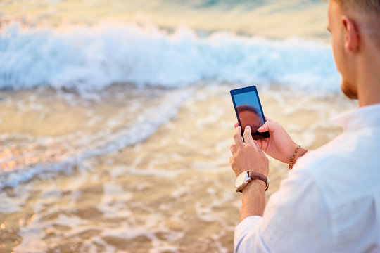 Vacation and technology. Young man using smartphone on the sea shore. Focus on screen.