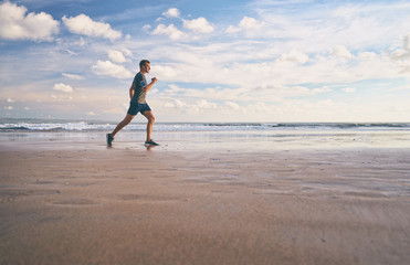 Enjoying sports lifestyle. Happy young man in headphones jogging on the sea shore.