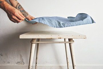 Man with tattooed arms puts a blue and white tablecloth over a small white old table with rust stains