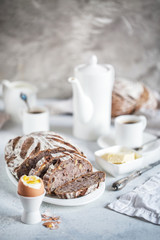 Artisan sourdough bread with rye flakes and walnuts in light breakfast composition with white porcelain, coffee, butter and hard-boiled egg