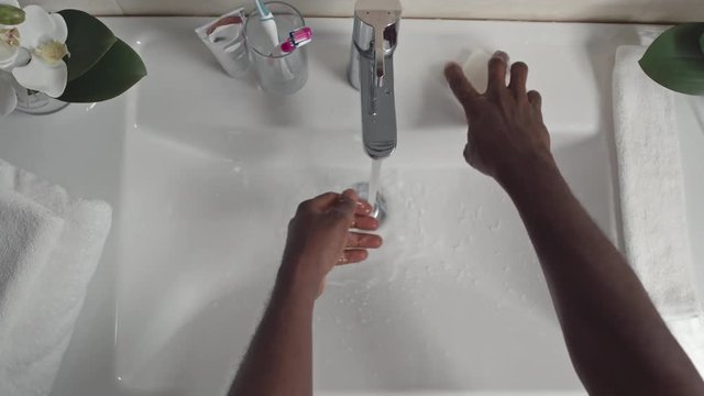 Directly above view of hands of African man washing hands with soap under water running from facet at sink and wiping them with white towel