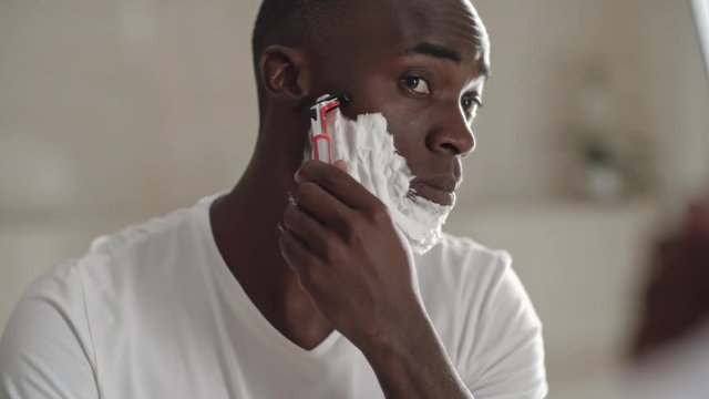 Closeup of African man gently moving razor on his cheek while shaving face with foam