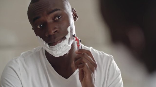 African man with creamy lather on his skin shaving face with razor