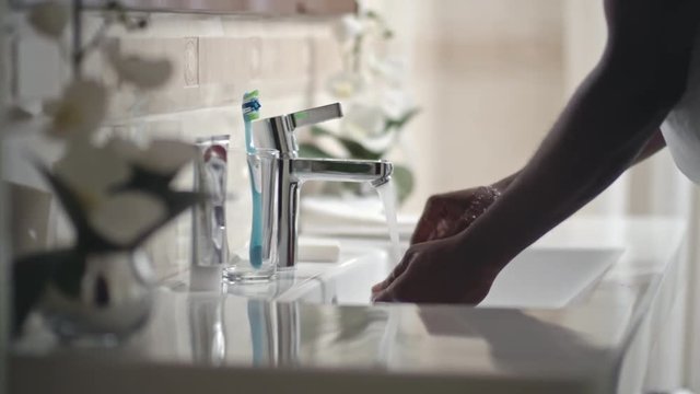 Tracking camera shot of hands of black man turning on water at sink and washing hands in bathroom