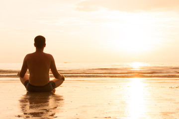 Fototapeta na wymiar Guy is sitting in lotus pose on sunset beach and meditating, enlightenment an zen concept