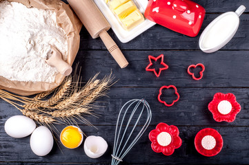 Ingredients for baking. Flour in paper bag, butter, milk, wheat, eggs and kitchen utensils - rolling pin and whisk on dark wooden background. Cooking cookies. Top view