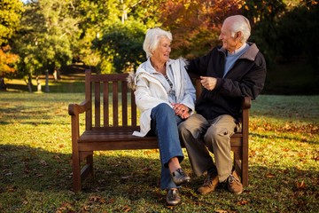  Elderly couple sitting on bench smiling at each other in park - Powered by Adobe