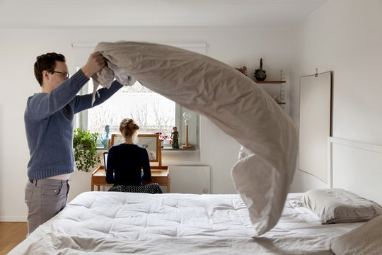 Man making bed while woman working at table in bedroom