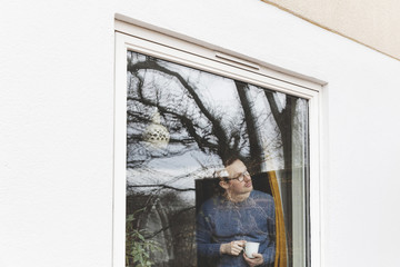 Man holding coffee cup looking through window while standing in house