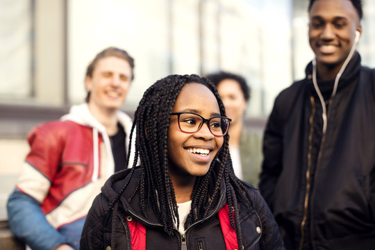 Smiling teenager with braided hair wearing eyeglasses standing against friends in city