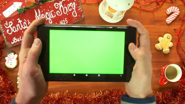 Man holding a tablet computer with green screen for easier screen replacement, background is a Christmas table top with cookies, coffee and other vintage decoration 