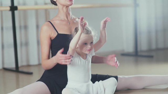 Stretching children. Little girl in the white and beautiful woman in black - teacher and pupil in dance school - doing stretching. A teacher at the ballet school teaches little dancer doing stretching