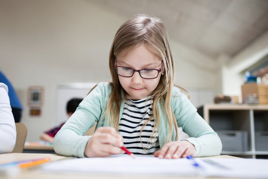 Concentrated girl wearing eyeglasses writing at classroom desk in school