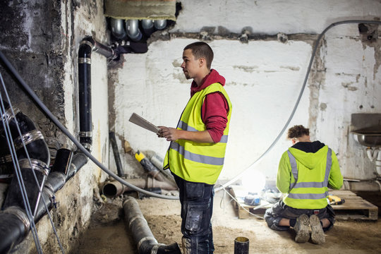 Side view of plumber analyzing document and looking at pipes while coworker working in background