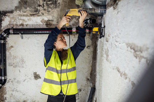 Confident woman drilling on wall above pipes at basement