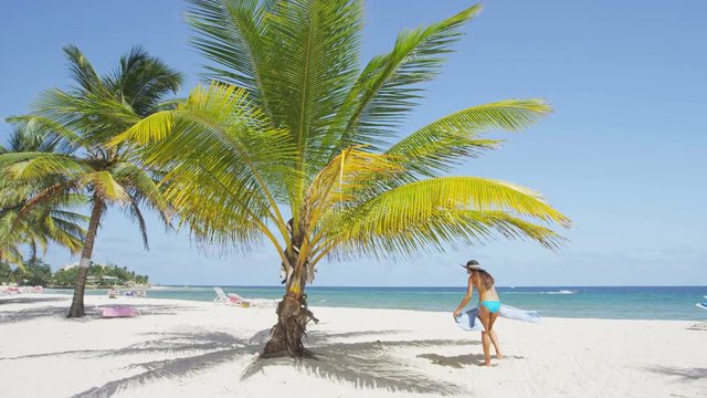 Woman on beach putting down beach towel for sunbathing under palm tree. Girl wearing bikini and sun hat relaxing on beach on Barbados, Caribbean. RED EPIC SLOW MOTION.