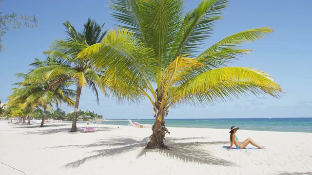 Beach travel sunbathing woman relaxing under sun in luxury retreat resort lying in sand. Woman tanning enjoying sun. Happy mixed race Asian Caucasian girl in bikini and sun hat. Barbados, Caribbean.