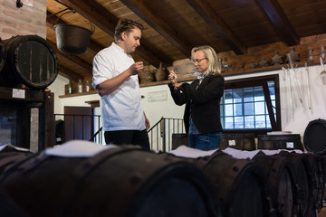 Chef and woman on balsamic condiment tasting in rustic cellar filled with wooden barrels