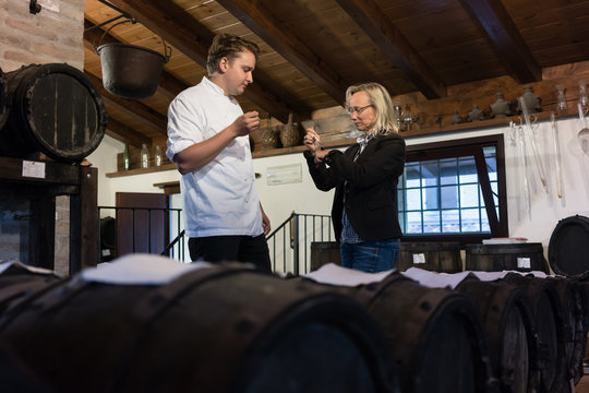 Chef and woman on balsamic condiment tasting in rustic cellar filled with wooden barrels
