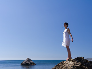 Naklejka na ściany i meble young girl in a white dress stand on the rocks above the sea, her hands like wings
