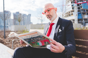 Middle-aged contemporary businessman sitting on a bench outdoor in the city reading newspaper -...