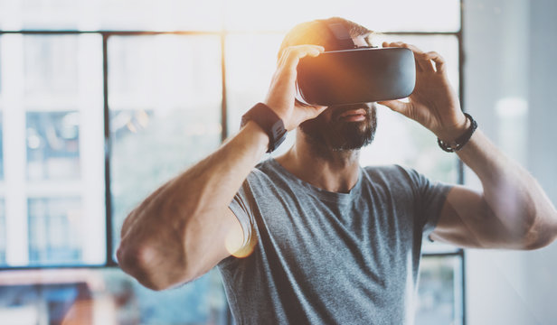 Attractive bearded man enjoyingvirtual reality glasses in modern interior design coworking studio.Home play concept.Smartphone use with VR goggles headset. Horizontal,flare effect,blurred background.