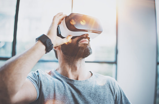 Attractive bearded man enjoyingvirtual reality glasses in modern interior design coworking studio.Home play concept.Smartphone use with VR goggles headset.Flare and sunny effect,blurred background.